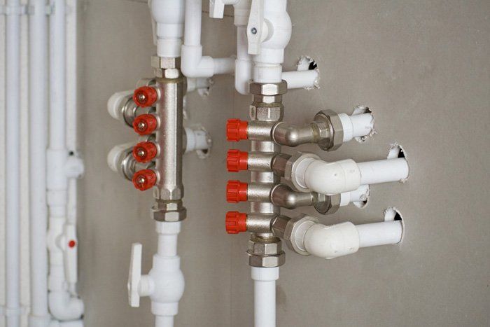 White and silver plumbing manifold with red valve handles against a light gray wall.