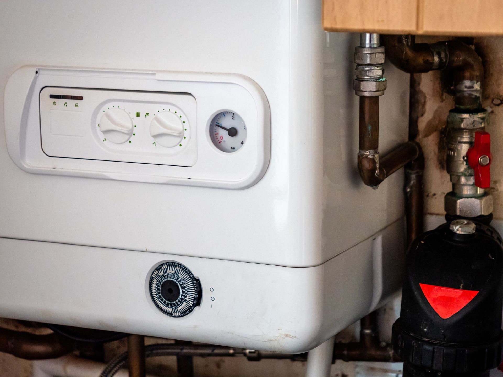 White wall-mounted gas boiler with dials and pipes, next to a black filter and red valve.