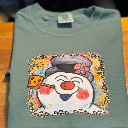 Green t-shirt with a smiling snowman design wearing a hat and smoking a pipe, framed by a leopard print border.
