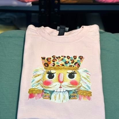 Pink t-shirt with a colorful embroidered nutcracker design. The nutcracker wears a leopard print crown and is against a green backdrop.