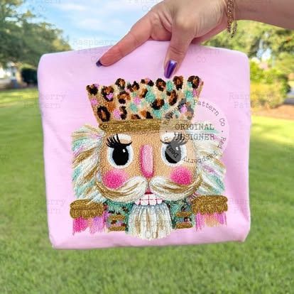 Pink shirt with embroidered Nutcracker wearing a leopard print crown, held by a hand.