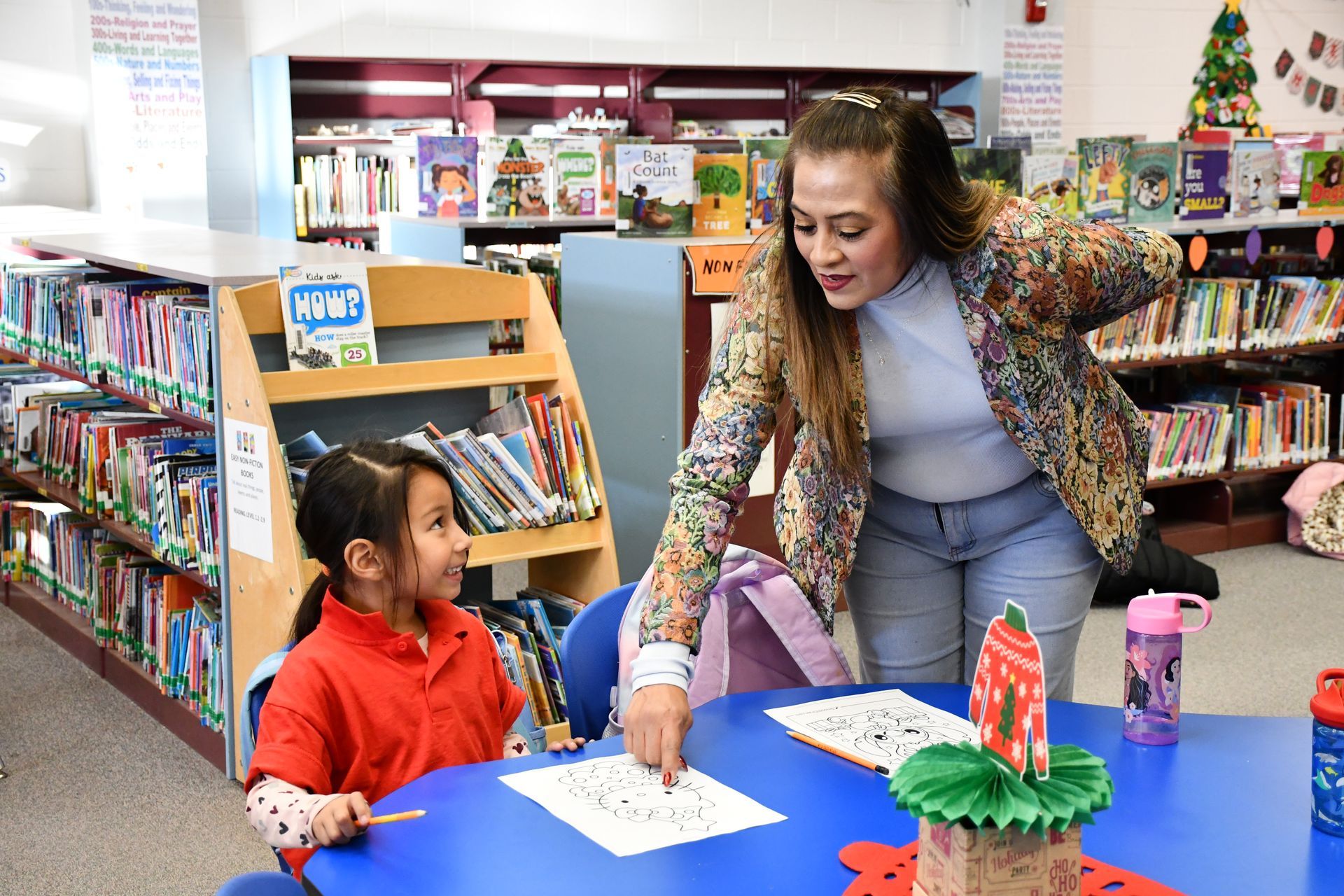 Anthony Elementary School student smiles as she looks at her teacher. The child is sitting at a desk while the teacher is standing and pointing to a paper on the desk.