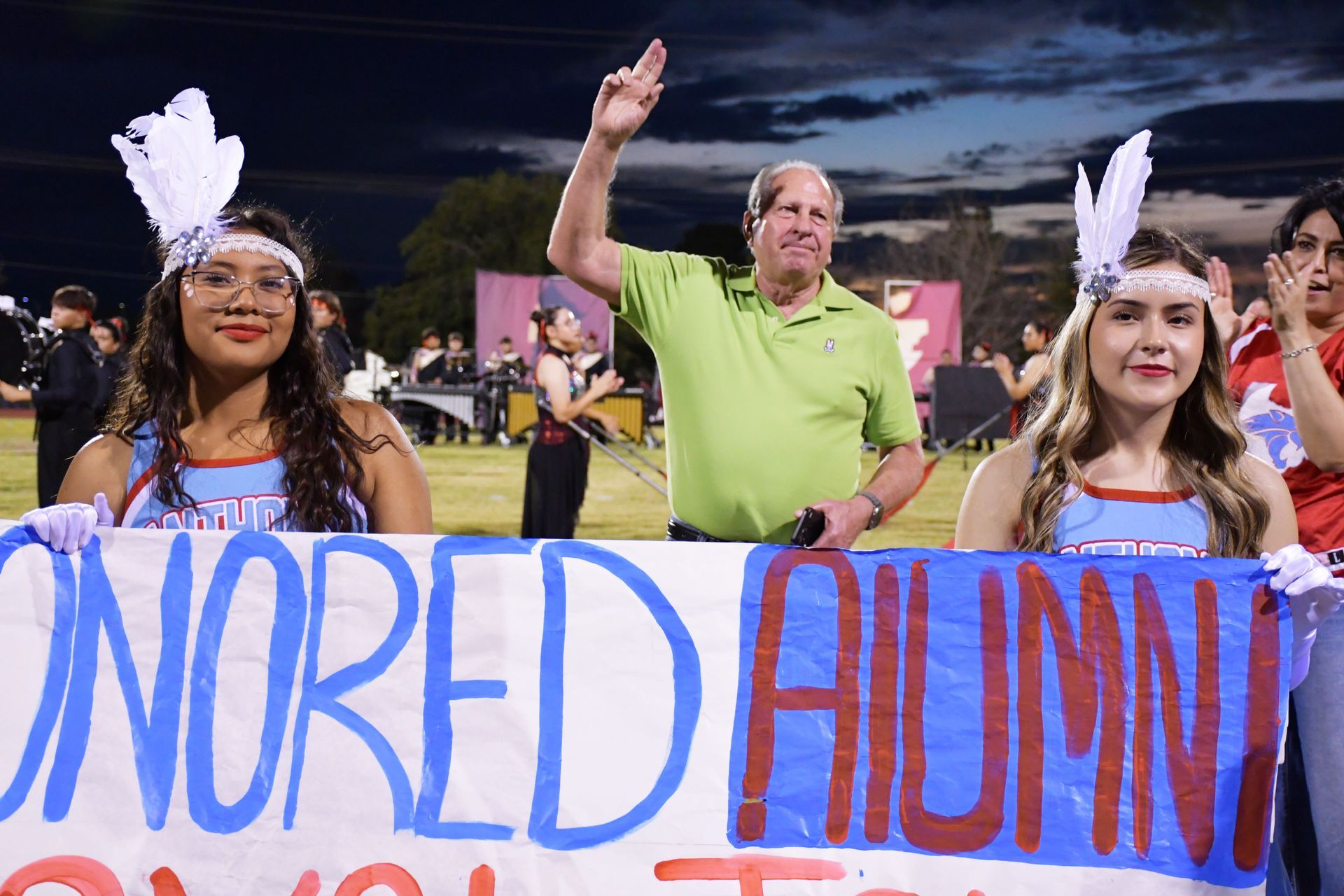 Man wearing green shirt salutes the audience from the football field while cheerleaders in front of him hold sign that says 