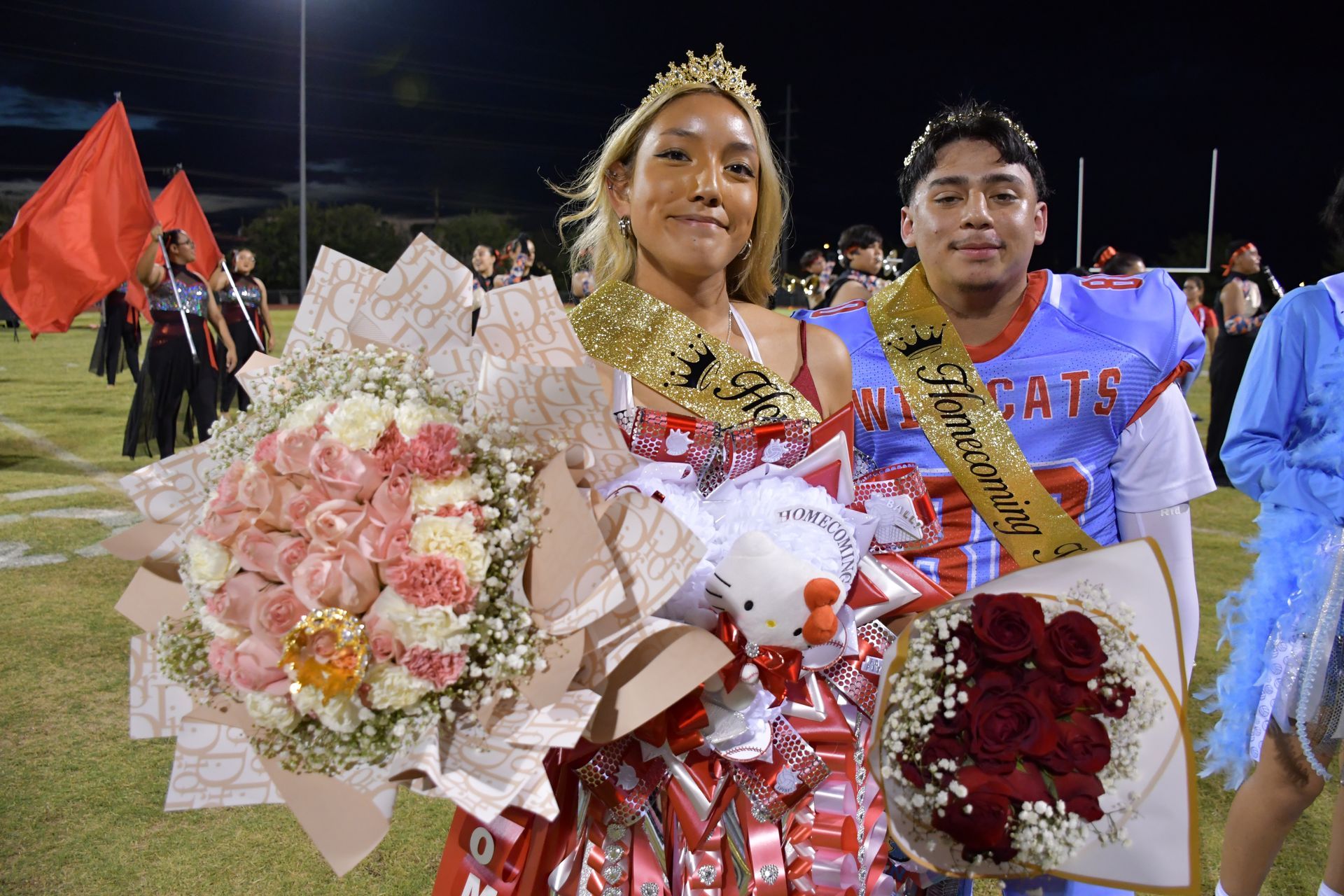 Female and male students stand on football field wearing homecoming mums and crowns for homecoming queen and king.