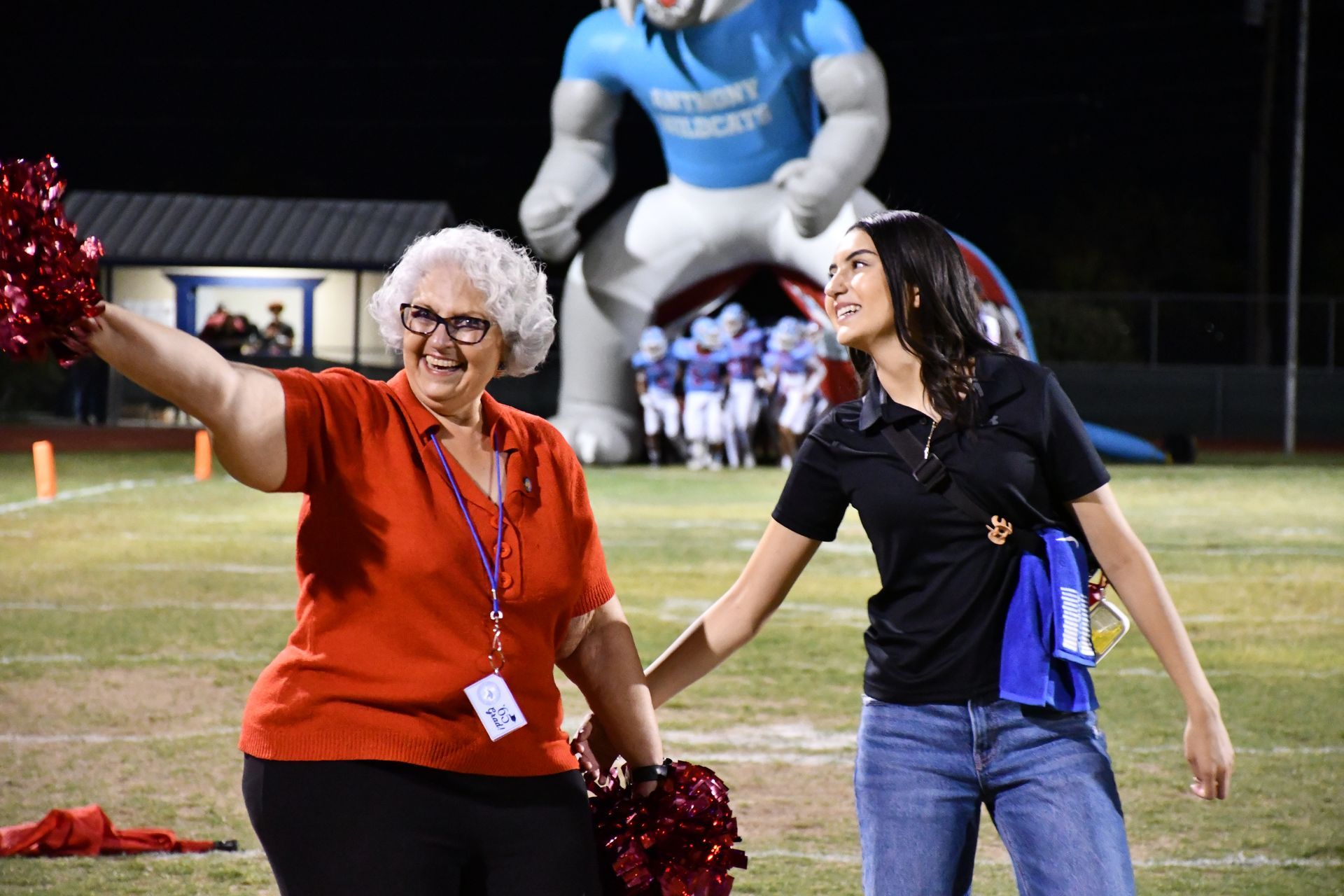 Woman in red shirt waves at crowd while holding a pom-pom. Student wearing a black shirt smiles and reaches for the woman.