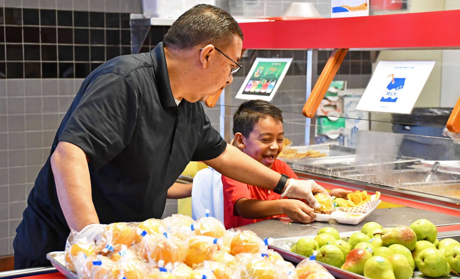 Child Nutrition Department employee gives an orange to a smiling child in the cafeteria line.