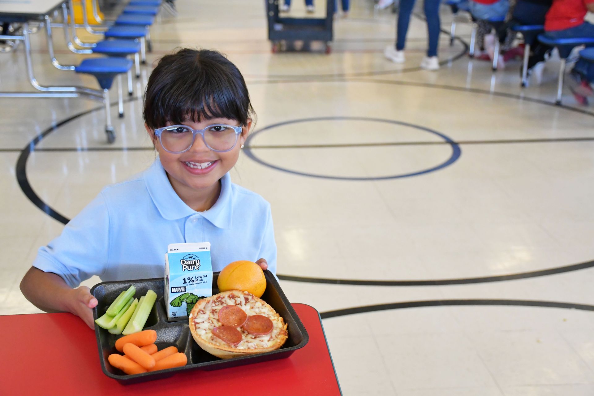 Child wearing light blue polo shirt and glasses holds food tray with pizza, carrots, celer sticks, an orange and a milk carton