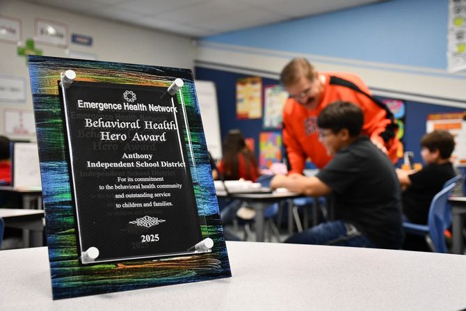 Behavioral Health Hero Award plaque sits on a desk in a classroom. In the background of the award, you can see a teacher assisting a student.
