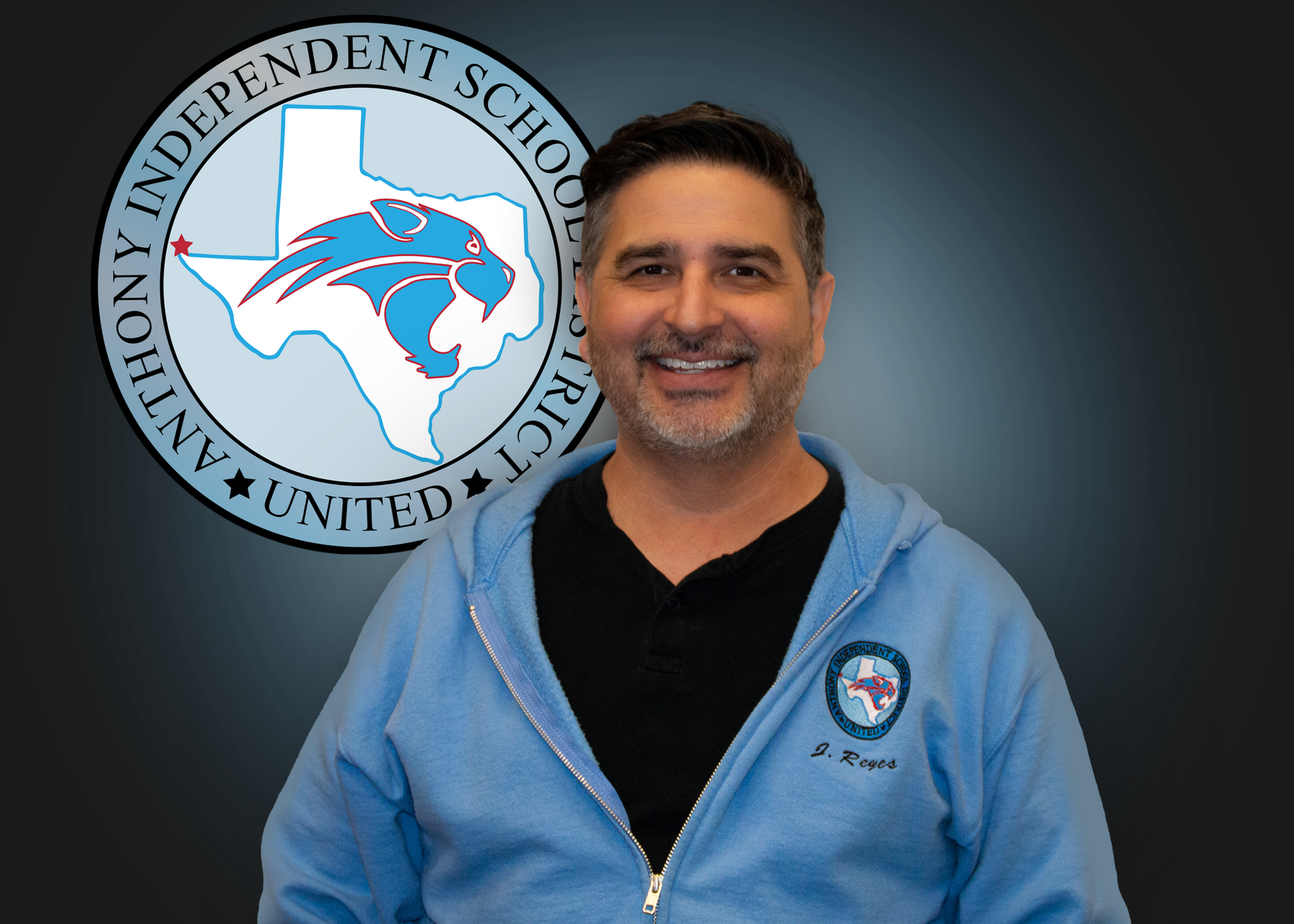 A man in a blue hoodie is smiling in front of an independent school district logo