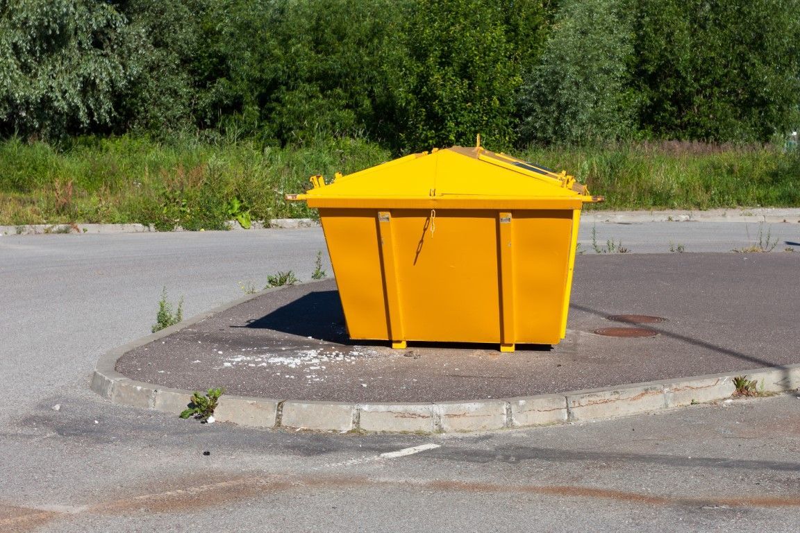 Yellow dumpster on concrete curb in a paved area, with trees in the background.