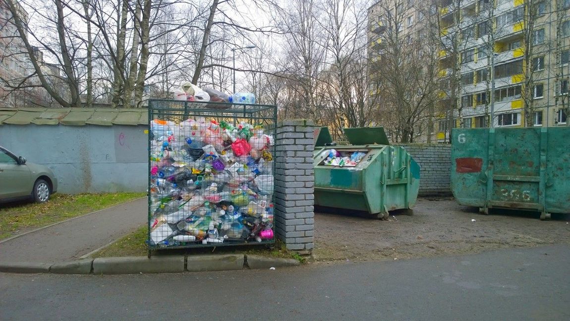 Overfilled recycling bin, garbage bins, and a building. Outdoors on a concrete area with a car.