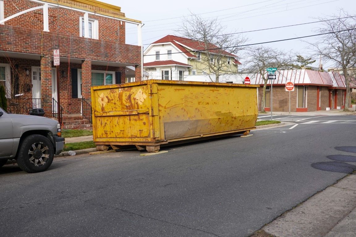 Yellow dumpster on street in front of buildings, with a pickup truck on the left.