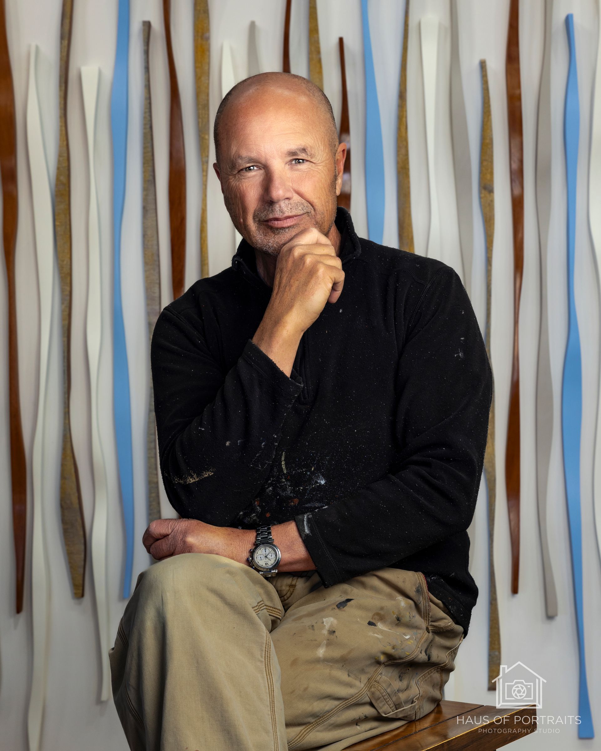 Person seated in black shirt before a wall of vertical wooden slats, resting chin on hand