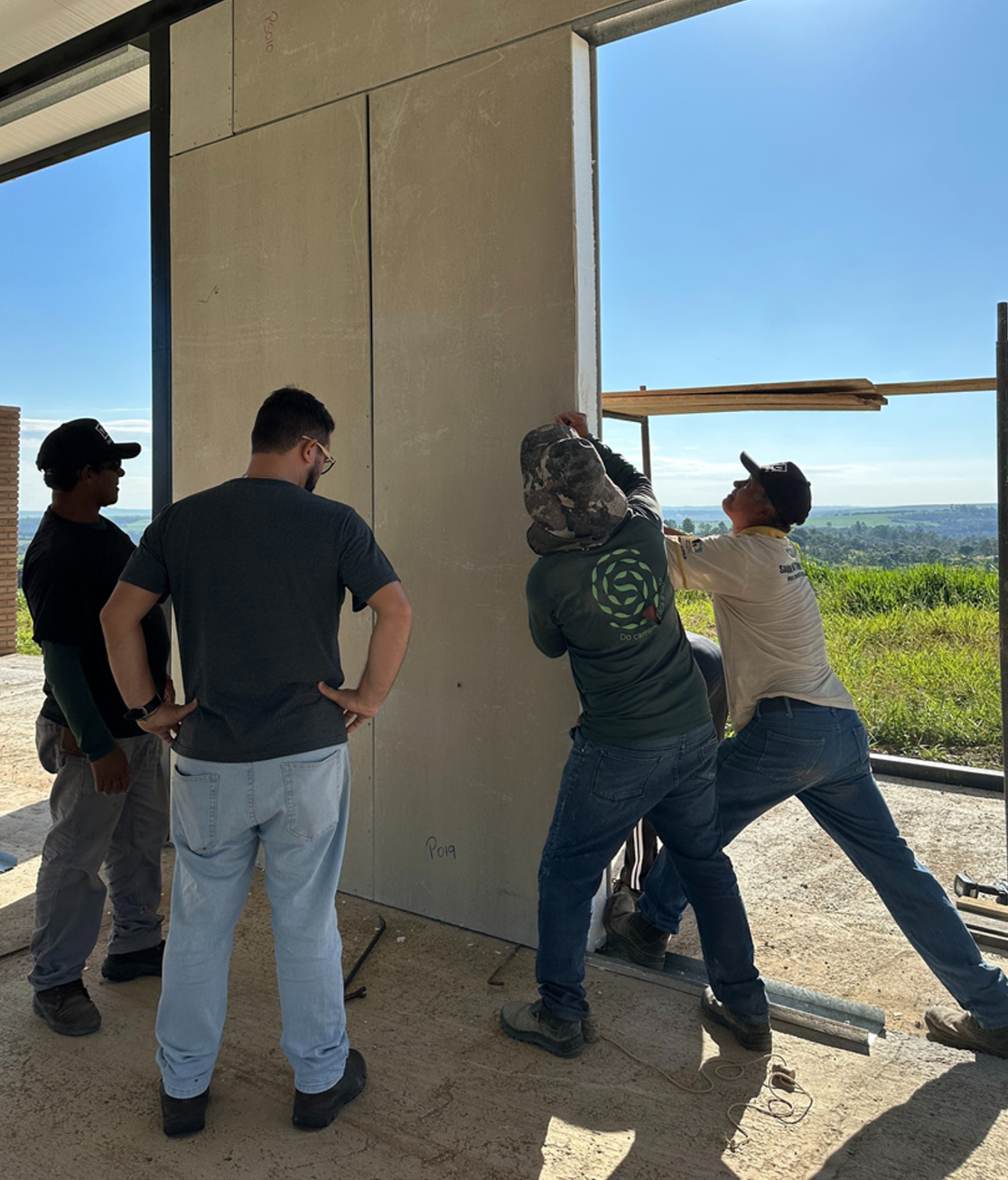 Operários da construção civil instalando painéis de parede ao ar livre. Eles estão perto de uma porta aberta com uma vista panorâmica ao fundo.