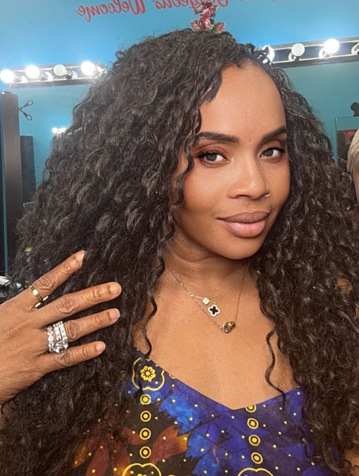 A close-up of a woman showcasing voluminous curly hair. She is smiling and posing with her hand placed near her hair, wearing multiple rings and necklaces. The backdrop is a bright blue salon wall with a welcoming sign in red above her.