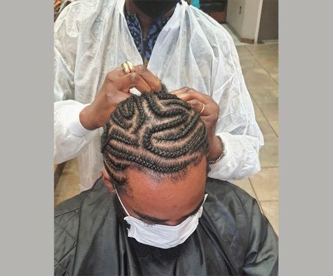 Person getting their hair braided in a unique pattern, with the braider's hands visible. Both individuals wear masks.