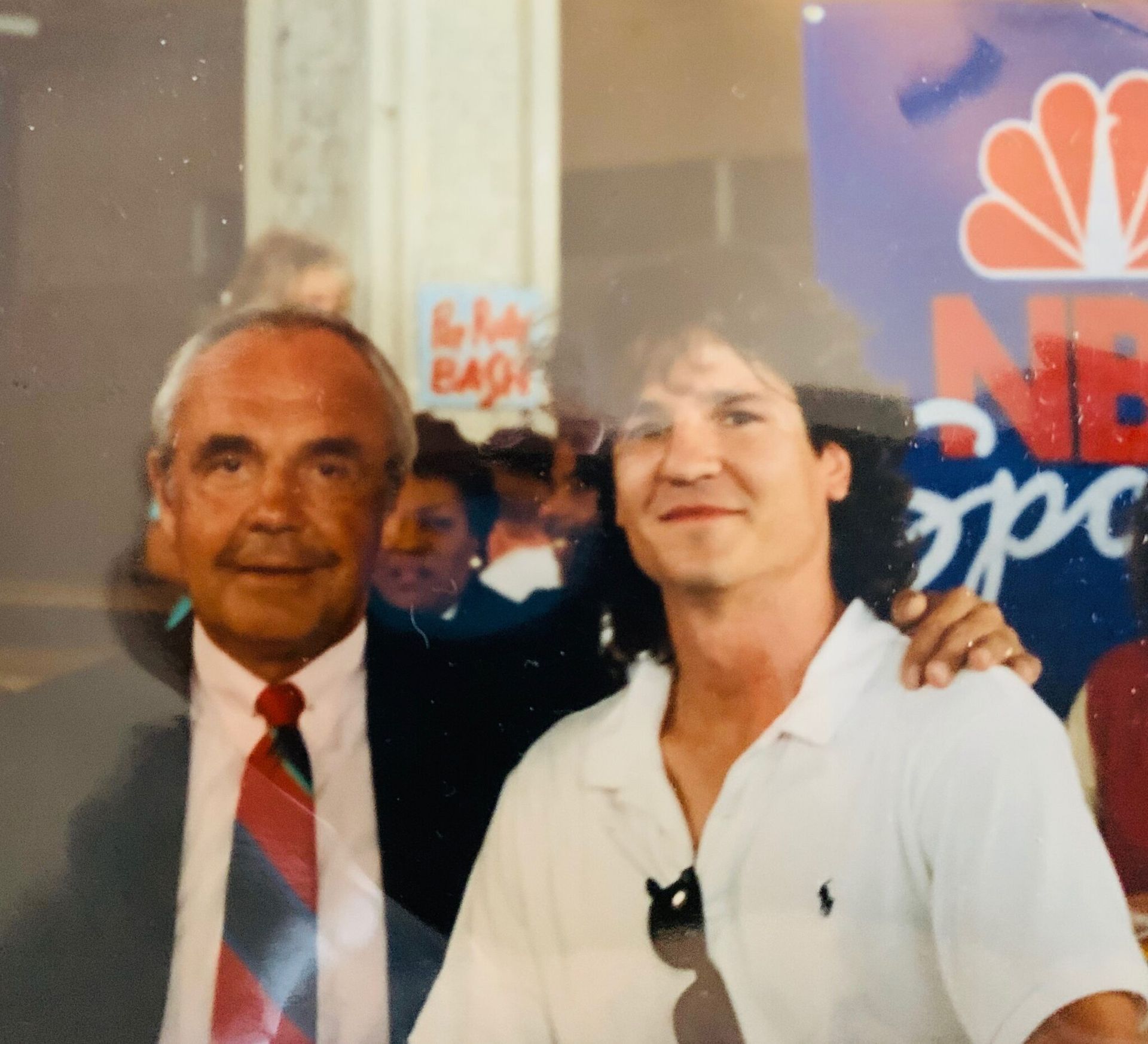 Two men are posing for a picture in front of a nbc sign