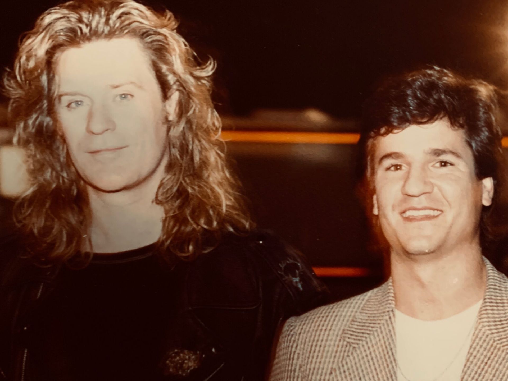 A man with long hair is standing next to another man in a suit.