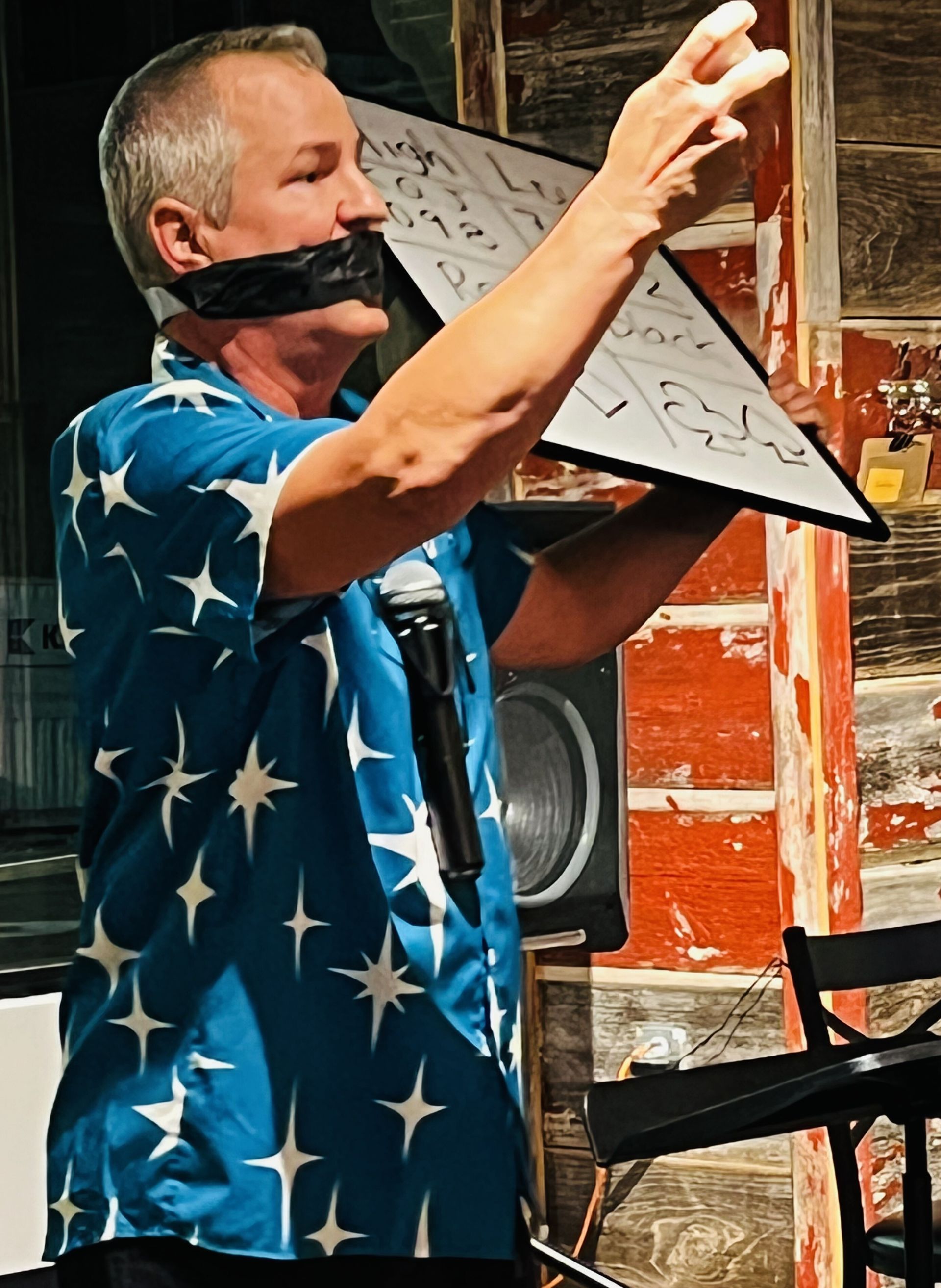 A man in a blue shirt with stars on it is holding a white board in front of his mouth.