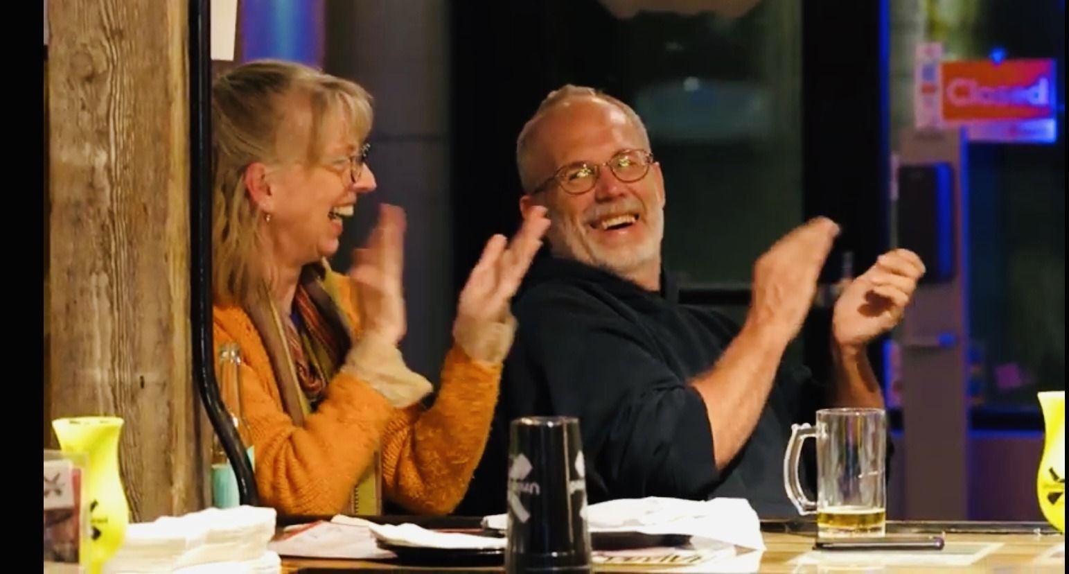 A man and a woman are sitting at a table clapping their hands.