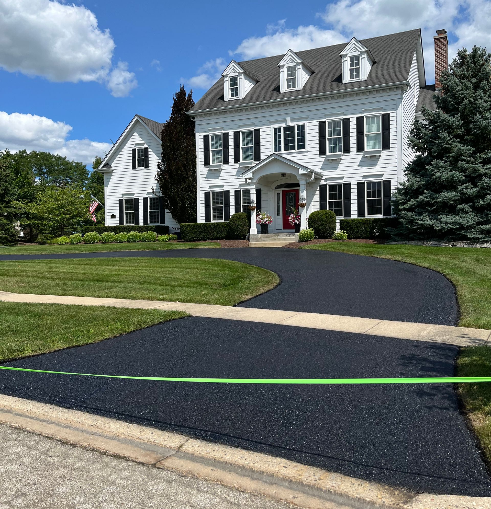 A large white two-story suburban home with a newly paved asphalt driveway and a bright green caution tape across the front.