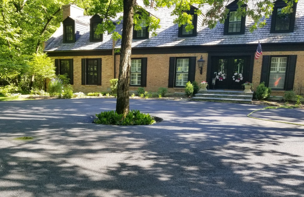 A two-story tan brick house with black shutters and trim, set behind a paved circular driveway featuring a centered tree.