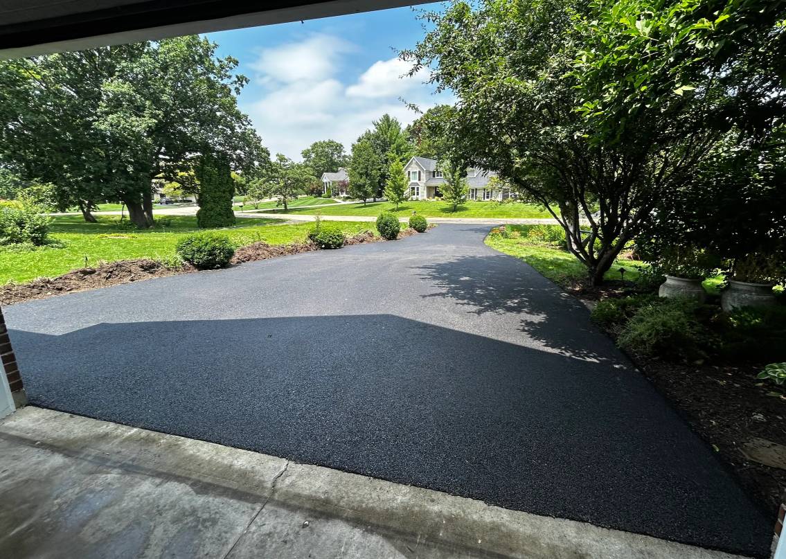 A fresh black asphalt driveway stretches toward a grassy yard with trees and a house in the distance.