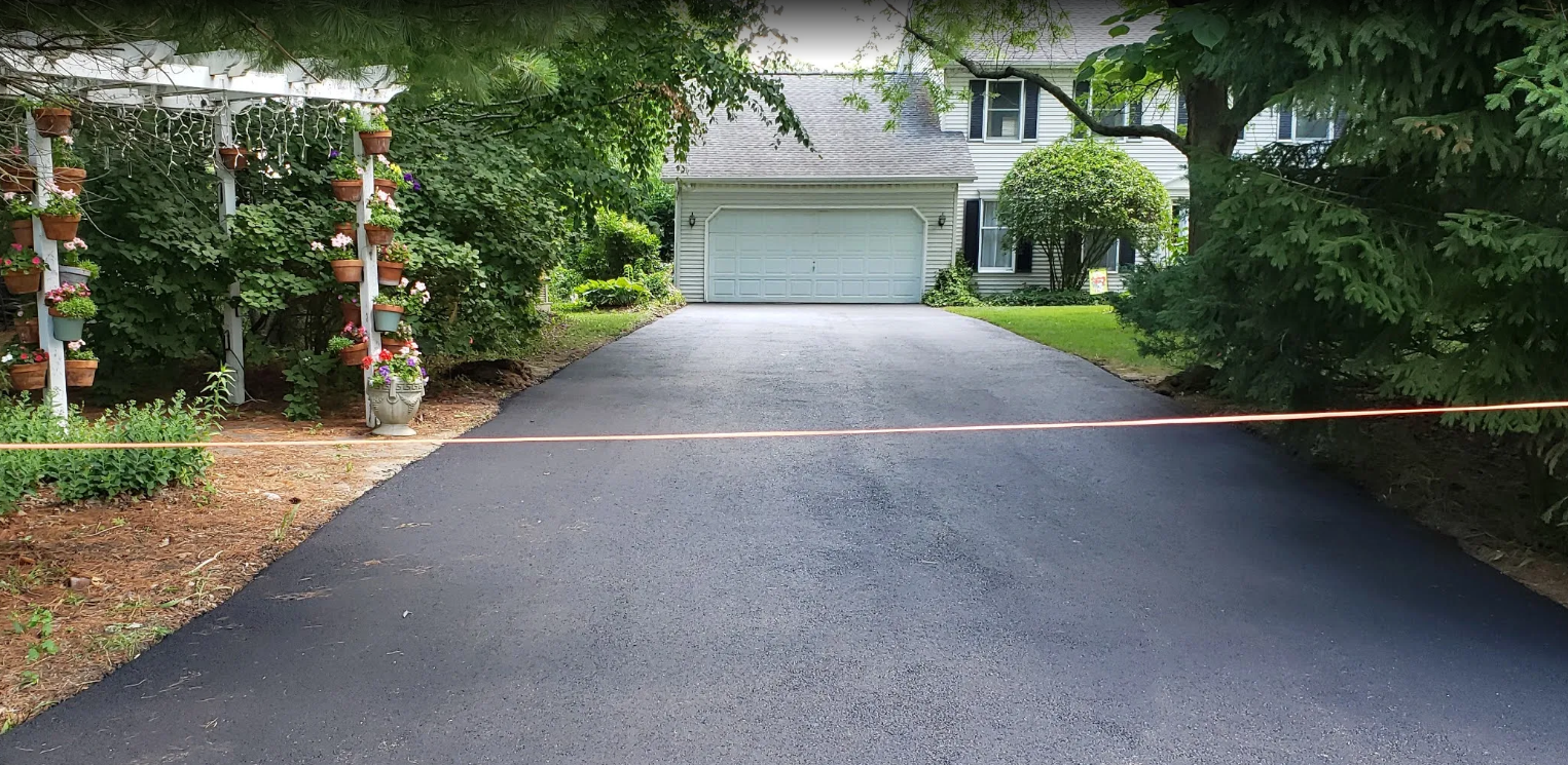 A fresh, dark asphalt driveway leads to a suburban garage, with trees and a plant trellis flanking the sides.
