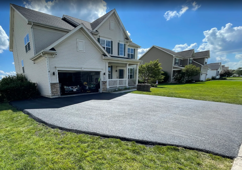 A suburban house with light-colored siding and a newly paved dark asphalt driveway under a blue, cloudy sky.