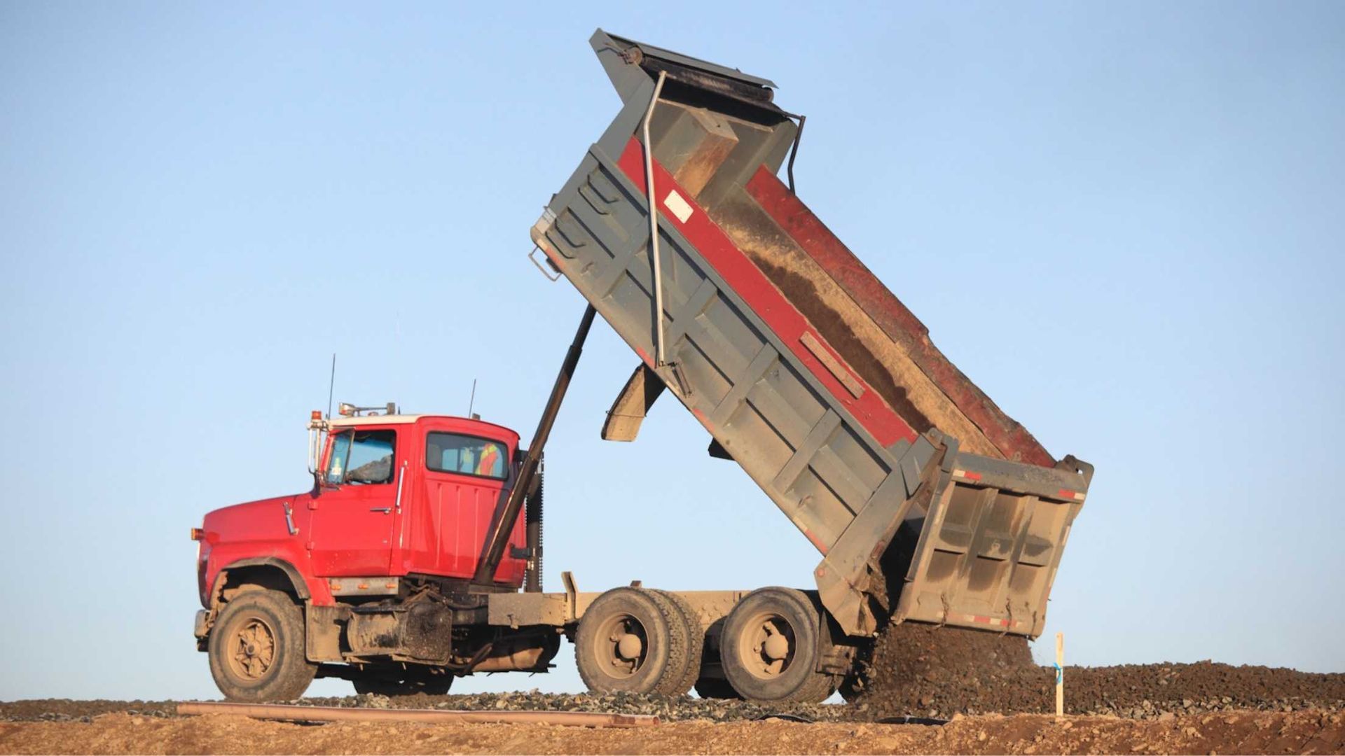 A red dump truck is dumping dirt on a dirt road.