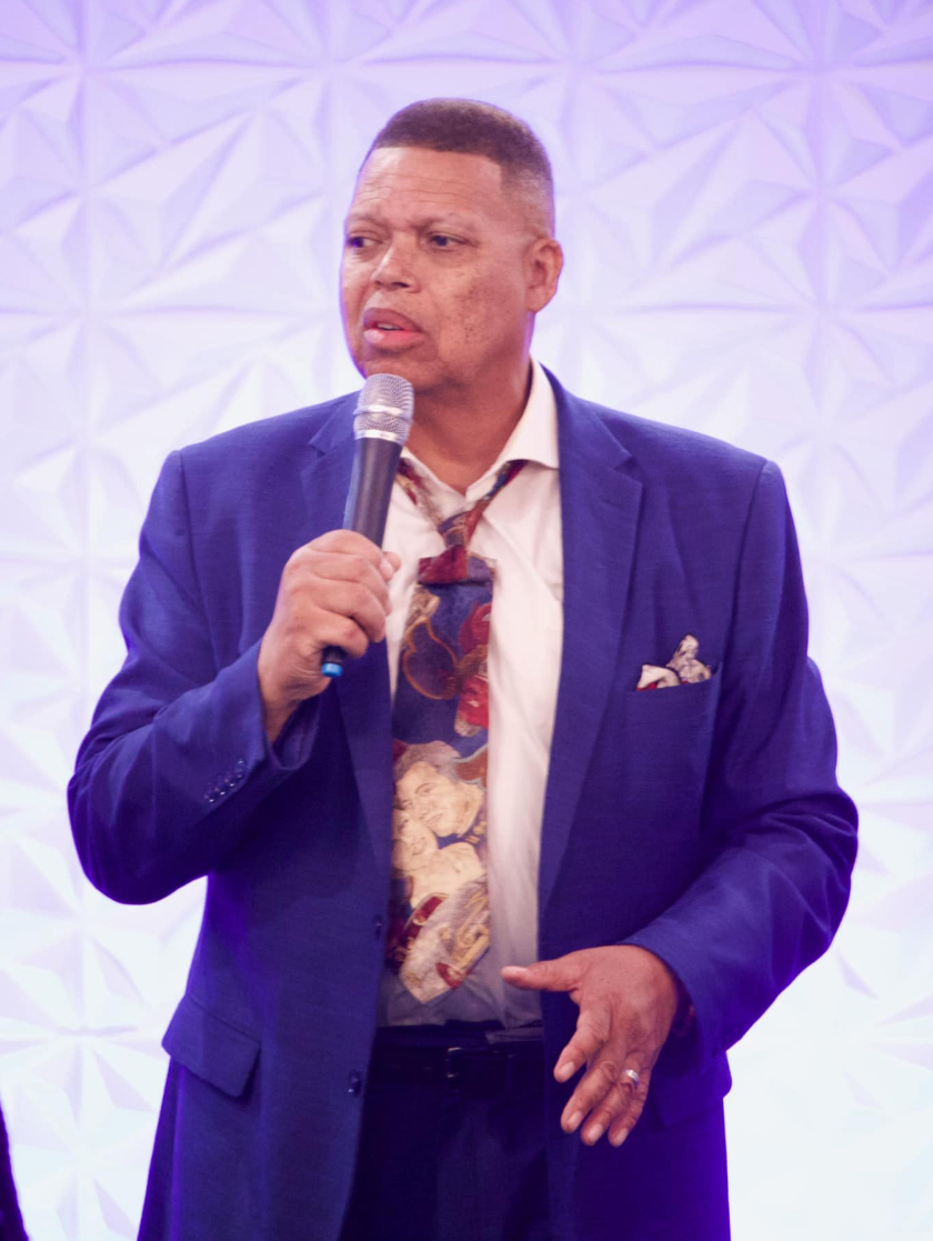 Man in blue suit speaks into a microphone; white wall background.