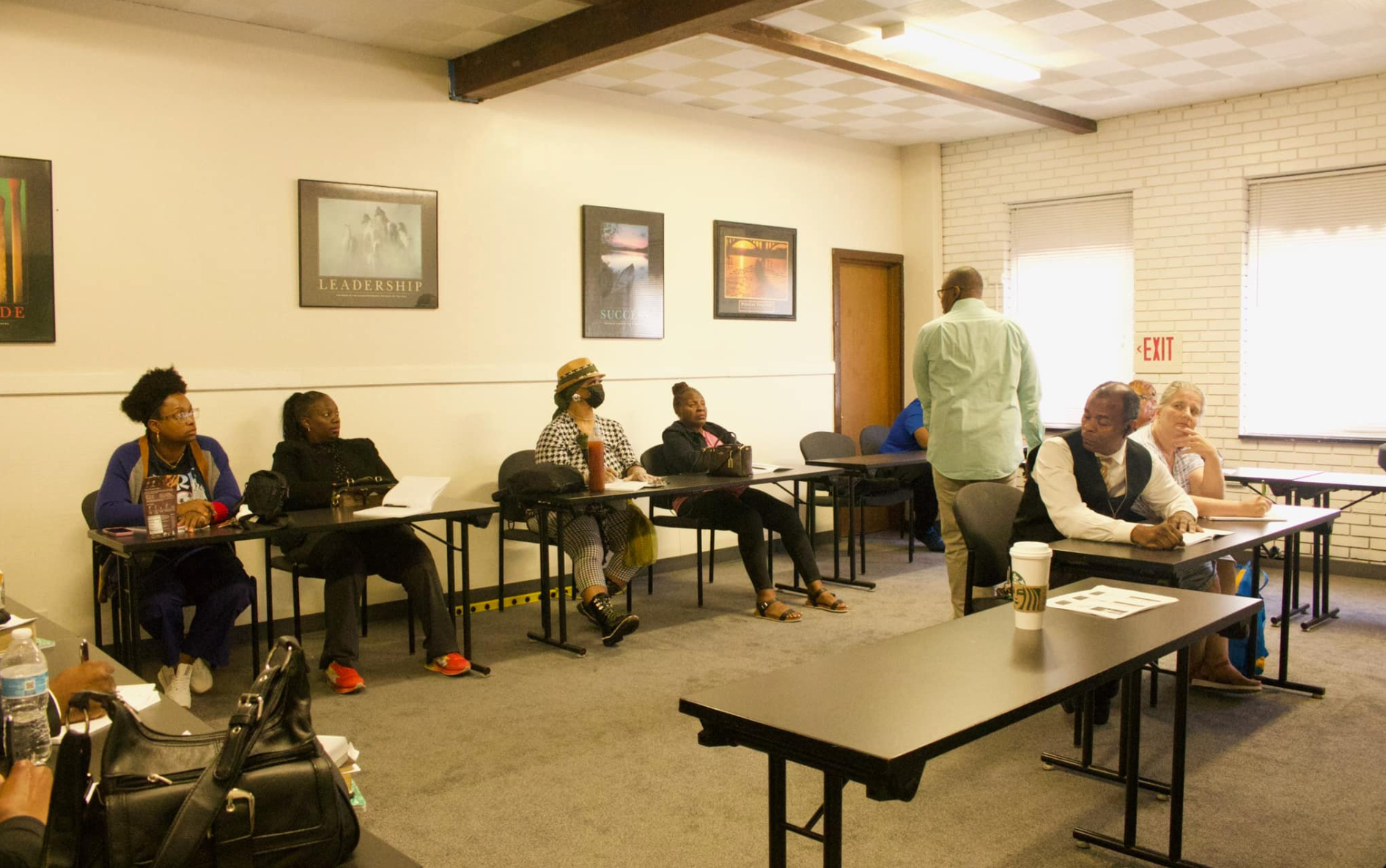 People seated at desks in a classroom setting; a man stands, possibly lecturing.