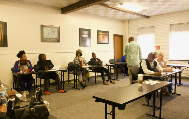 People seated at desks in a classroom setting; a man stands, possibly lecturing.