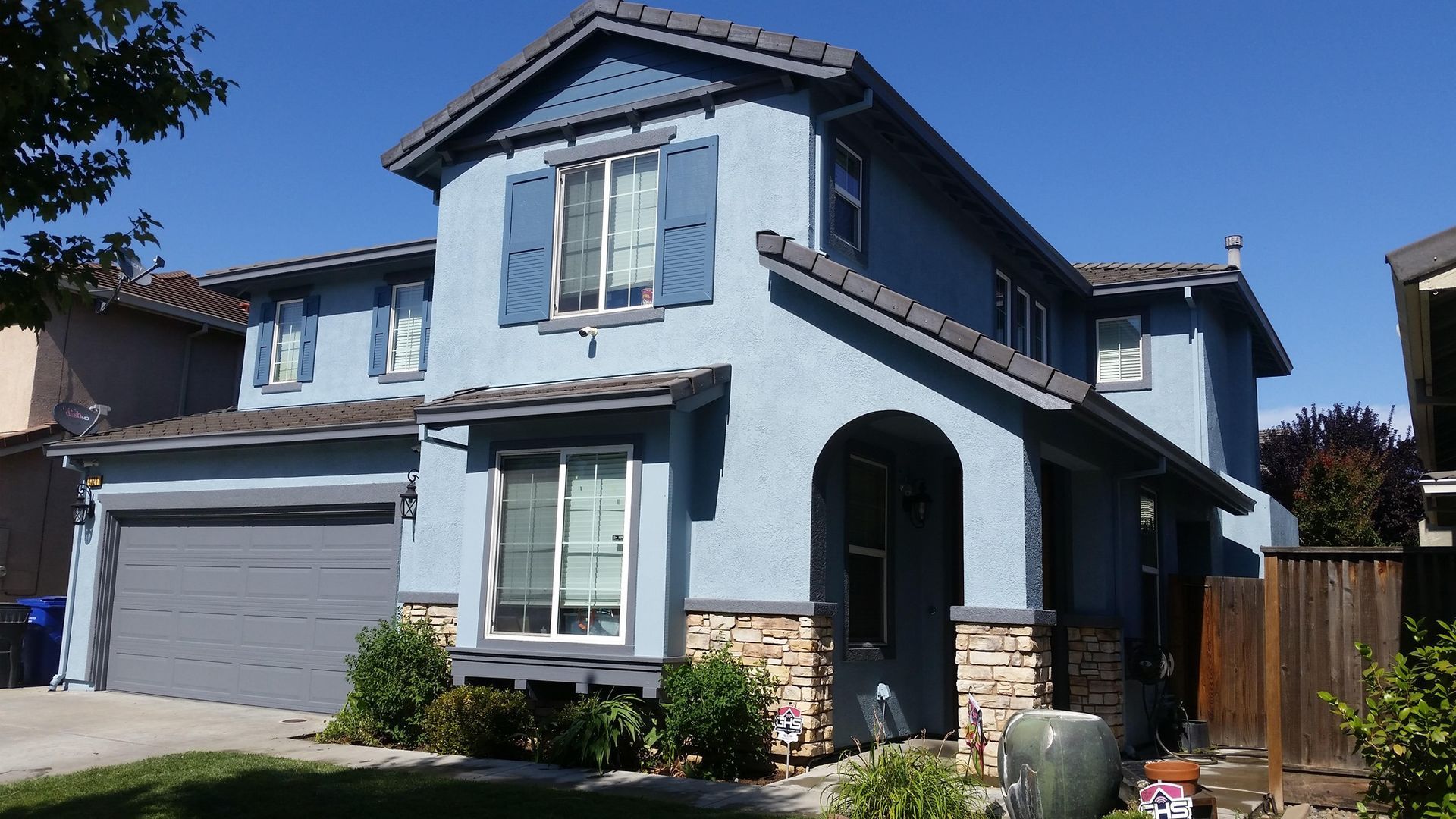 Two-story blue house with gray roof and shutters, stone accents, and a gray garage door on a sunny day.