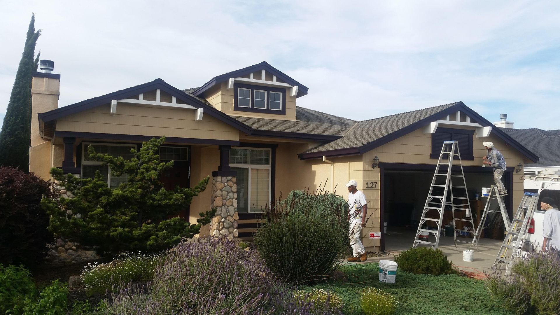 House being painted; beige walls, brown trim, painters on ladders, cloudy sky.