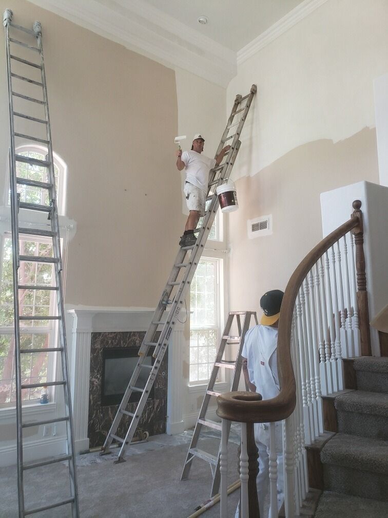Painter on a tall ladder painting a high wall. Another worker on a shorter ladder nearby. Interior shot.