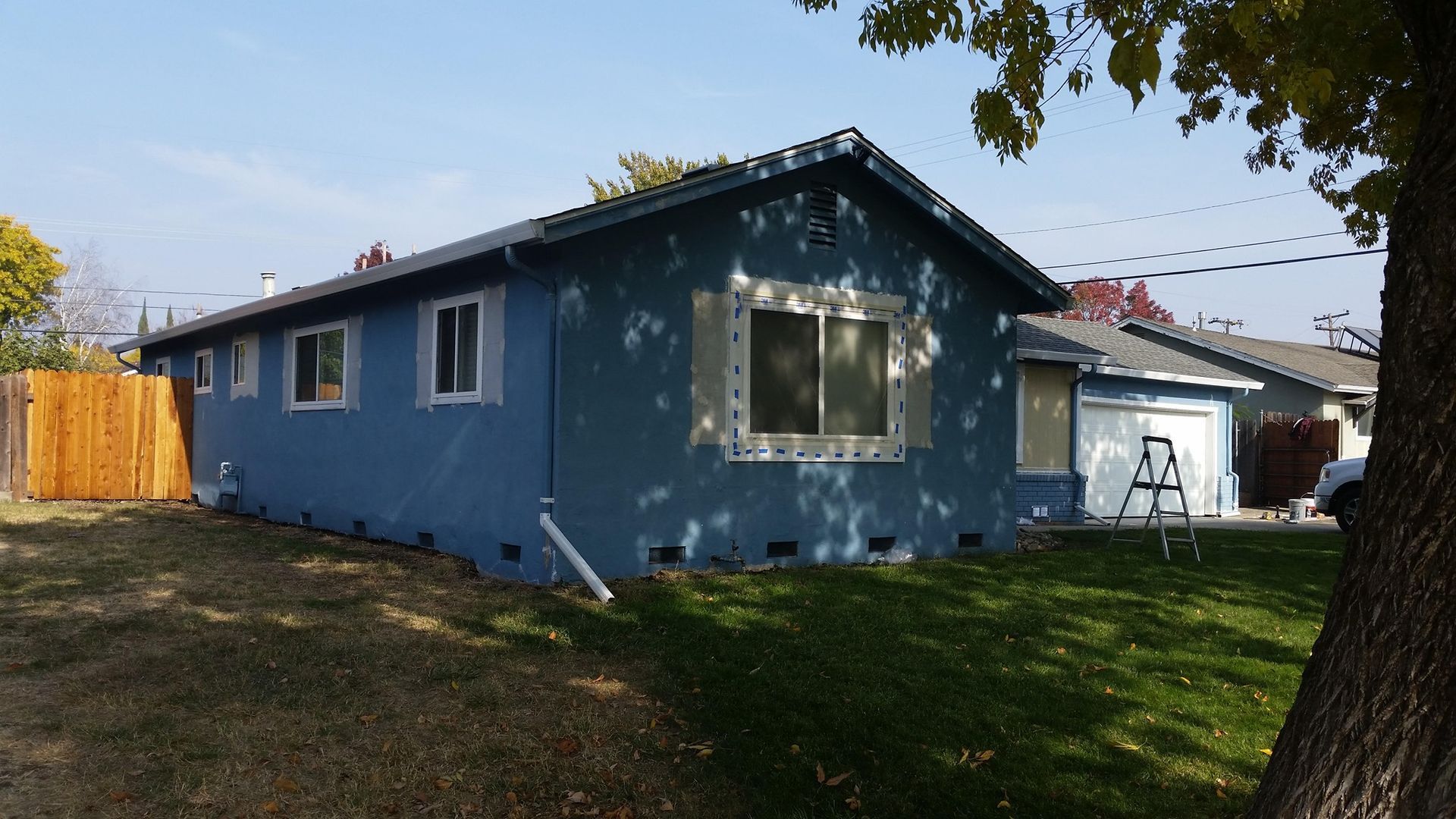 Blue house with white window trim, brown fence, green grass, and a tree on a sunny day.