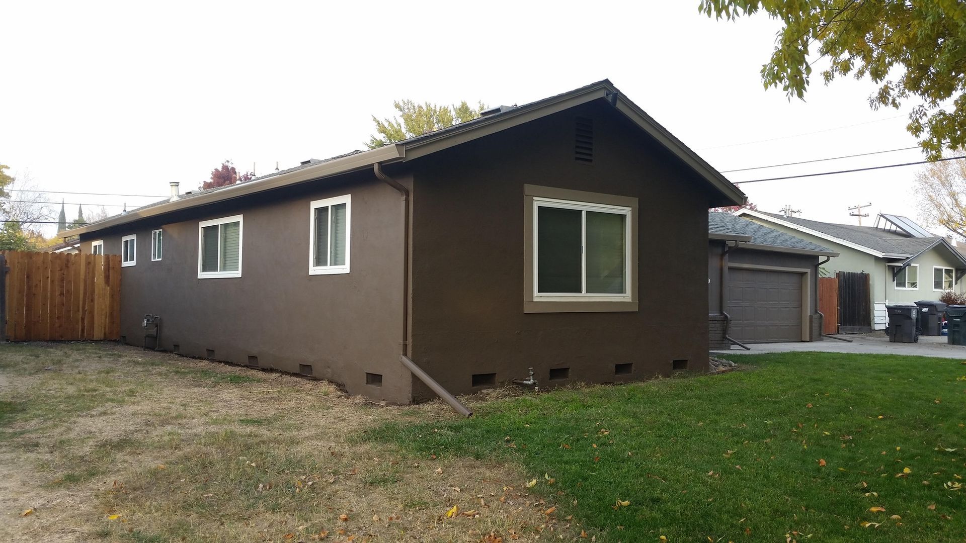 Brown house with white-framed windows. Wooden fence on the left, garage and lawn on the right. Cloudy sky.