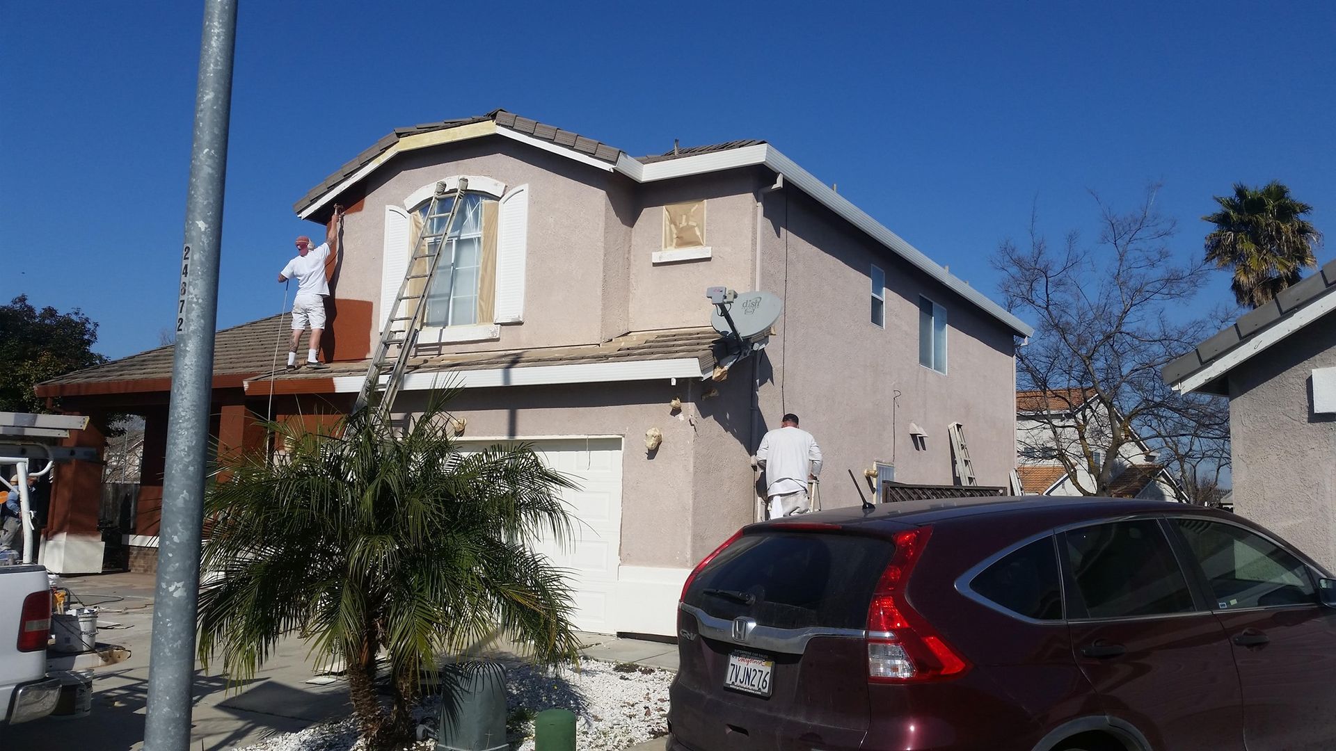 Two-story house being painted by workers; red car parked in front; clear blue sky.