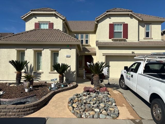 Two-story beige house with a tan garage door, red shutters, and a cloudy sky.