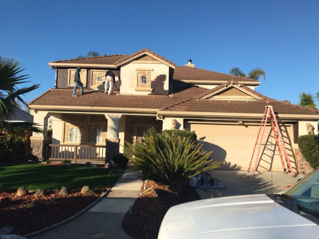 Roofers on a two-story house, working on the roof under a clear blue sky. A ladder leans against the house.