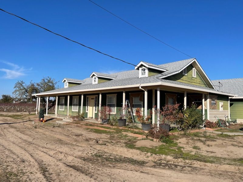 Green farmhouse with porch under a clear blue sky, surrounded by dirt and plants.