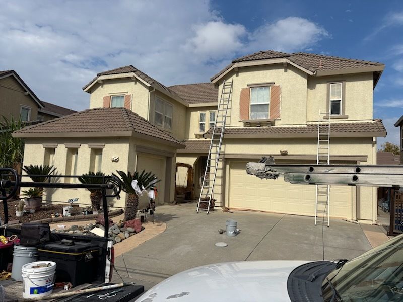 Backyard view of a white house with a dark roof, chimney, and patio. Plants, furniture, and a blue sky are present.