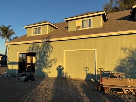Yellow house with gray roof, blue chairs, and wooden bench on a sunny day.