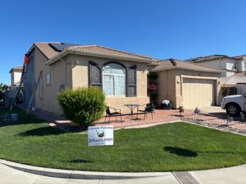 Backyard view of a white house with a dark roof, chimney, and patio. Plants, furniture, and a blue sky are present.