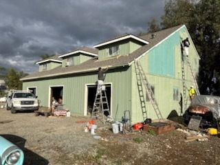 Back of a one-story house with a gray roof and tan siding, surrounded by dry grass and a blue sky.