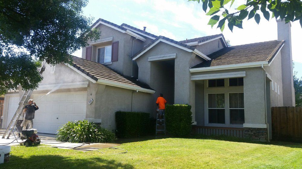 House being painted. Two workers on ladders, one in orange shirt. Green lawn, blue sky.