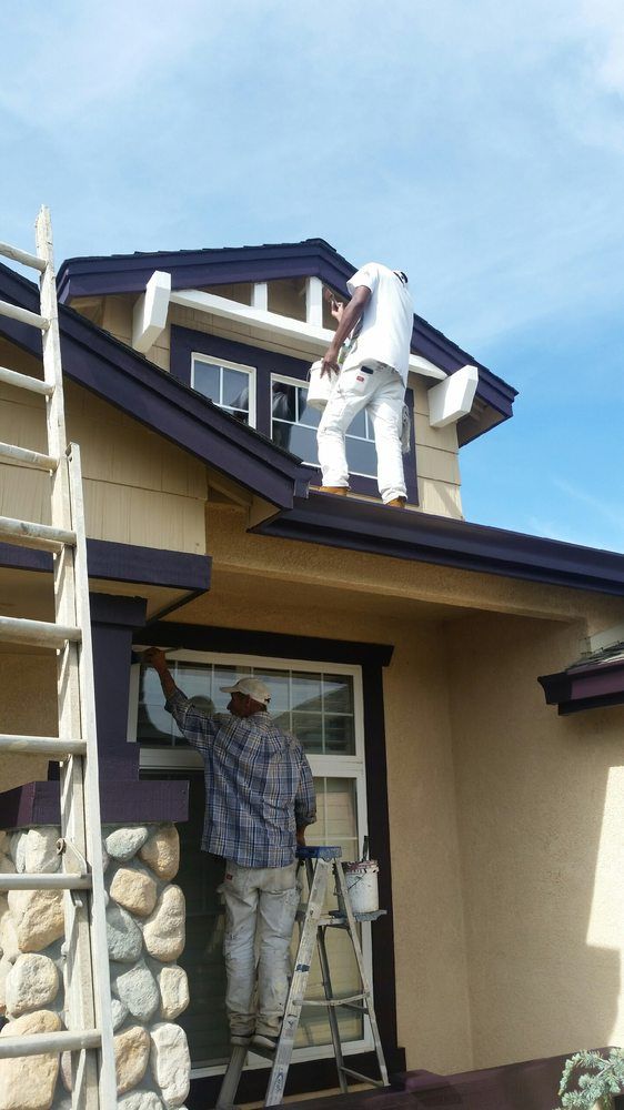 Two painters on ladders painting a house exterior. Dark purple trim against tan siding, blue sky.