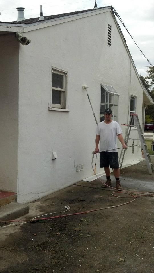 Man paints white house exterior with roller, standing outside on a cloudy day.