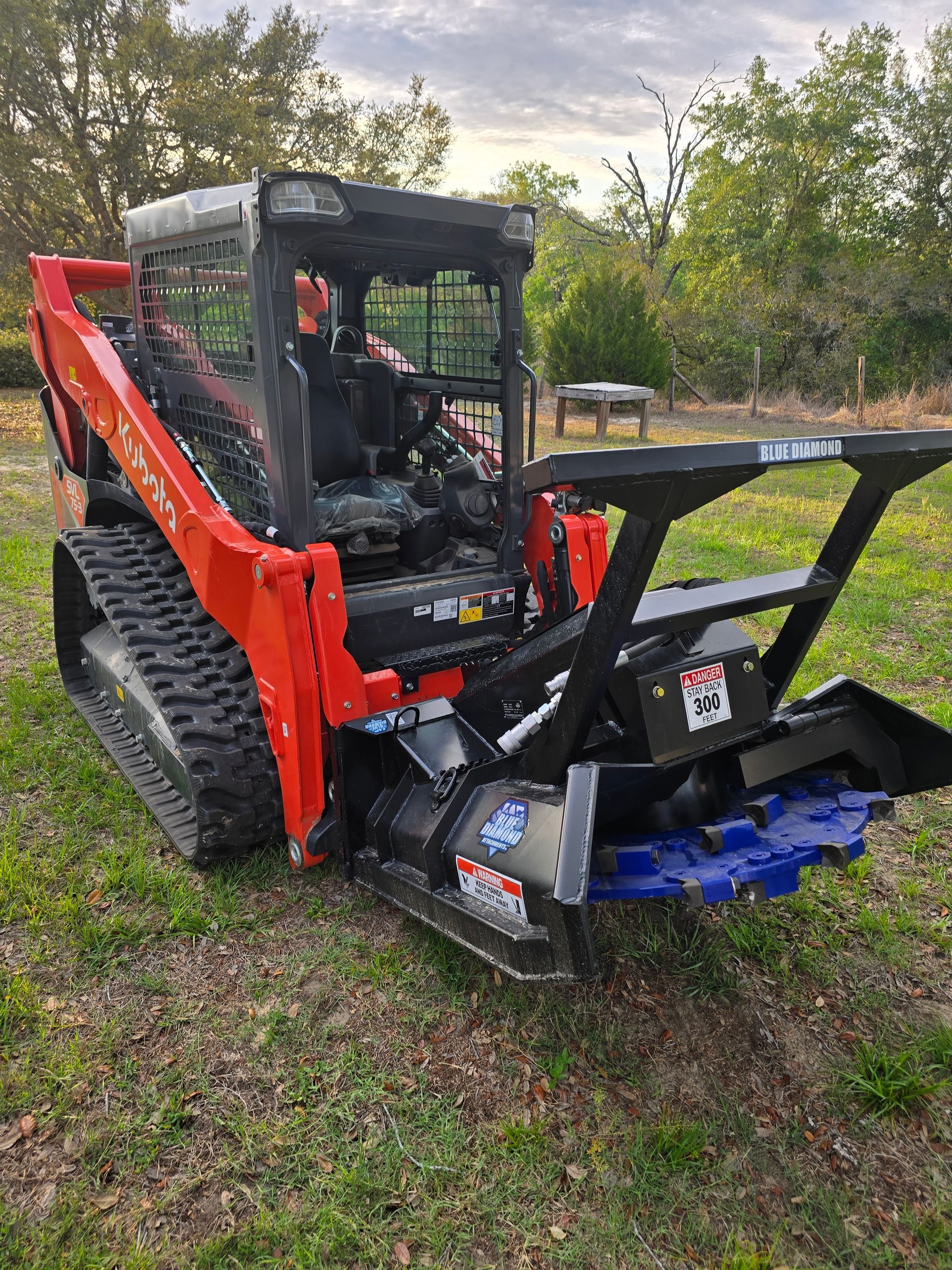 An orange and black track loader with a forestry mulcher attachment parked on a grassy field.